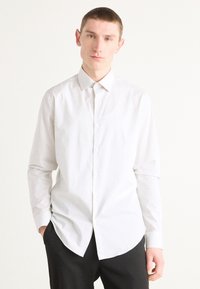 Camisa blanca de manga larga con botones, hecha de una tela ligera, que presenta un cuello clásico y un dobladillo curvado. Llevada con pantalones negros.