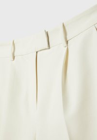 Gros plan de la ceinture d’un pantalon couleur crème avec passants de ceinture et détails plissés sur le devant.