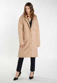 Beige gewatteerde lange jas met een capuchon, drukknoopsluiting aan de voorkant en een ritsdetail in de zak. Vlakke textuur en een lichte glans. Draag over zwarte broek.