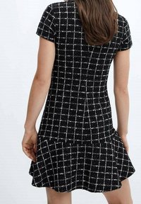 Mujer con cabello castaño vistiendo un vestido negro de manga corta con un patrón de cuadrícula blanca y dobladillo acampanado, vista de espaldas.