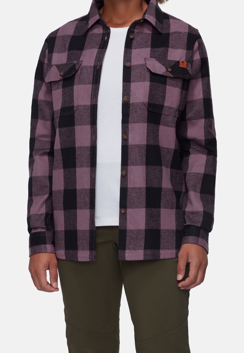 Personne portant une chemise à carreaux violette et noire à boutons par-dessus une chemise blanche et un pantalon vert olive, montrée des épaules aux hanches.