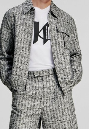 Man draagt grijs-zwarte tweedjas en broek met wit Karl Lagerfeld logo t-shirt, handen in jaszakken, neutrale achtergrond.