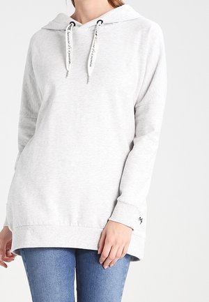 Femme portant un sweat à capuche gris clair à manches longues avec poche avant et cordons blancs, associé à un jean bleu sur un fond uni.