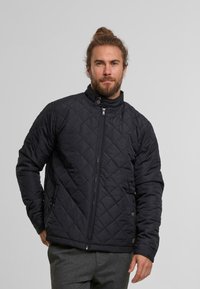 Pikowana czarna kurtka z zapięciem na zamek i wysokim kołnierzem. Posiada kieszenie boczne i fakturowany materiał. Nosi się ją na szarych spodniach.