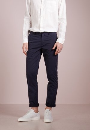 Homme portant une chemise blanche boutonnée, un pantalon bleu marine retroussé et des baskets blanches, debout devant un fond beige uni.