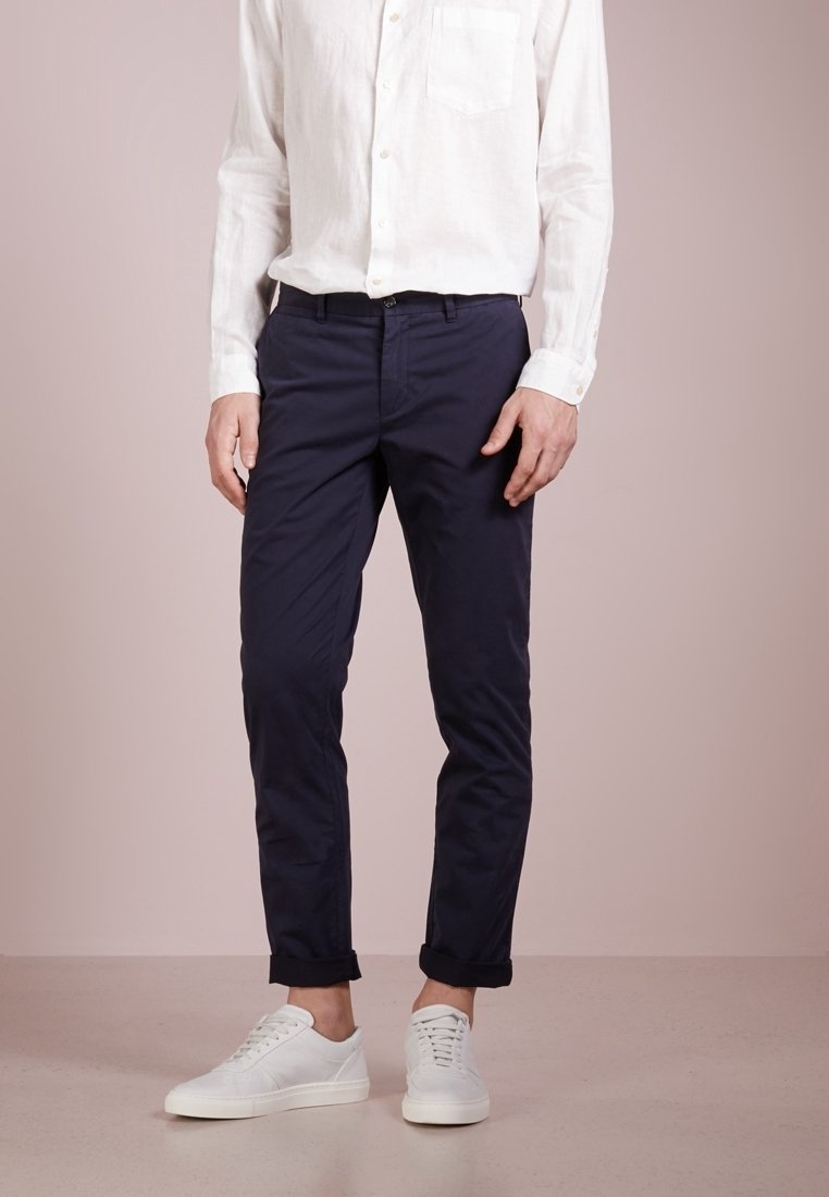Homme portant une chemise blanche boutonnée, un pantalon bleu marine retroussé et des baskets blanches, debout devant un fond beige uni.