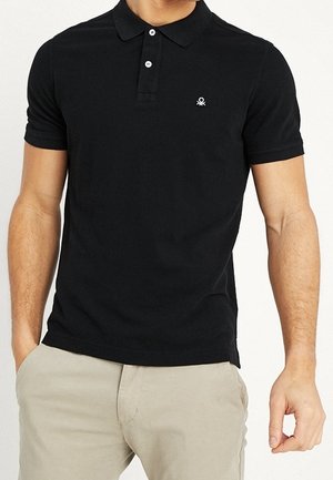 Mand iført en sort kortærmet polo med to hvide knapper og et lille hvidt logo på venstre bryst, kombineret med beige bukser.