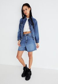 Veste en denim bleue au design court, associée à un short en denim bleu. Des bottines à lacets noires complètent la tenue. Texture lisse partout.