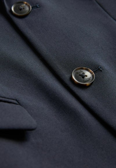 Marinblå kavaj med texturerad tyg, försedda med två runda, metallknappar med ingraverad detalj och en sned ficka med lock.