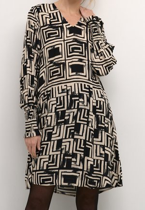 Kvinde iført en beige kjole med sort geometrisk firkantet mønster og lange pufærmer, stående mod en ensfarvet baggrund.