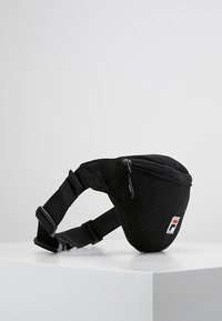 Borsa a vita nera in rete con cerniera, dotata di cinturino regolabile e piccolo logo rettangolare sul davanti, esposta su una superficie bianca.