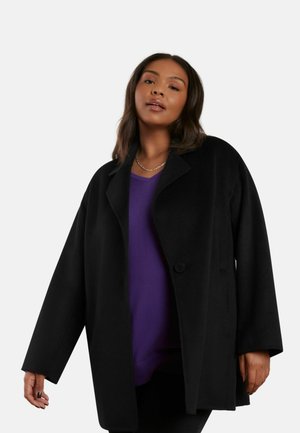 Cappotto oversize nero realizzato in materiale morbido, con colletto a punta, chiusura a singolo bottone e maniche strutturate. Indossato sopra un top viola.