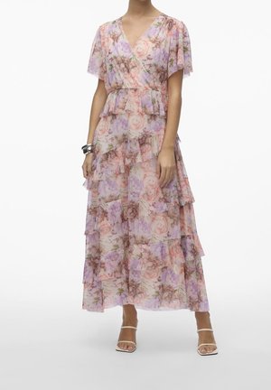 Kvinde iført en blomstret maxikjole med lag og korte, flagrende ærmer og V-hals, kombineret med hvide remsandaler og et sølvarmbånd.