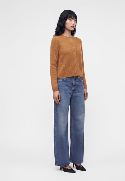 Femme debout portant un cardigan marron boutonné, un jean bleu à jambes larges et des talons pointus noirs sur un fond clair uni.