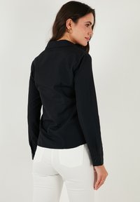 Black long-sleeve shirt with a collared neck. Fitted design, smooth texture, and buttoned cuffs. Worn with white fitted pants.