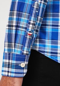 Manica di una camicia a quadri blu e bianchi con polsino beige e un piccolo bottone decorativo rosso, bianco e nero sulla patta della manica.