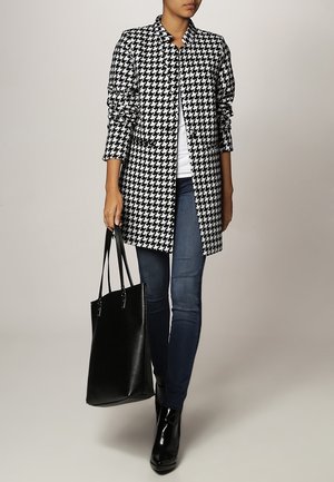 Hahnentritt-Mantel in Schwarz und Weiß, mit langen Ärmeln, ohne Knöpfe vorne, kombiniert mit blauen Jeans und einer schwarzen Tragetasche. Glänzende Knöchelstiefel.