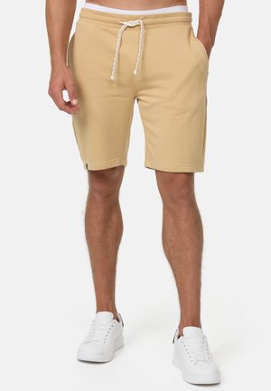 Mann trägt beige Kordelzug-Shorts und weiße Sneakers, steht mit einer Hand in der Tasche vor einem schlichten Hintergrund.