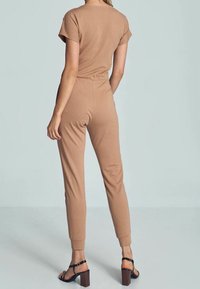 Combinaison beige avec manches courtes, taille élastique, jambes ajustées et poignets côtelés. Portée avec des sandales à talons marron, vue de dos.
