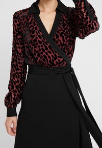 Rød og sort leopardprint indpakningsbluse med sort krave, lange ærmer og taljebind; blød tekstur og glat finish.