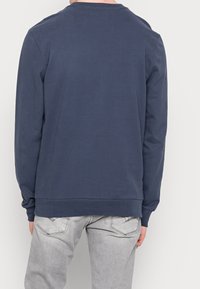 Muž oblečený v námořnicky modré mikině s dlouhým rukávem a světle šedých džínách stojí čelem k bílému pozadí, zády ke kameře.