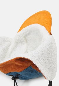 Chapeau avec un extérieur en velours côtelé orange texturé et une doublure en polaire blanche duveteuse. Dispose d'une base bleue et marron avec un cordon de serrage.