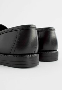 Chaussures en cuir noir avec une texture lisse, un talon bas et des coutures visibles. Le design est élégant avec un dos arrondi et une légère courbure.