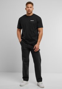 Sort bomulds T-shirt med rund hals og korte ærmer, der har et hvidt "DEFSHOP" logo på brystet; sat sammen med mørke jeans og hvide sneakers.