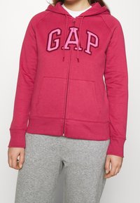 Donna con una felpa con zip di un rosa scuro GAP, con stringhe sul cappuccio e pantaloni della tuta grigio chiaro, in piedi davanti a uno sfondo neutro.