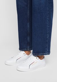 Białe sneakersy z gładką skórzaną powierzchnią, z złotymi detalami, czerwonymi i granatowymi akcentami oraz nadrukowanym logo. Połączone z niebieskimi dżinsami.