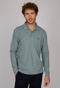 Polo à manches longues en tissu doux de couleur bleu clair, avec un col, un logo discret sur la poitrine et des poignets côtelés. Un ajustement classique avec un minimum de détails.