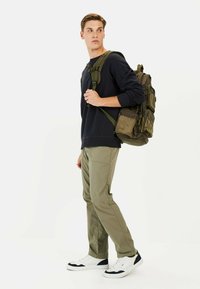 Groene stoffen rugzak met meerdere zakken, gedragen door een persoon in een zwarte trui, groene broek en witte sneakers, staande tegen een witte achtergrond.