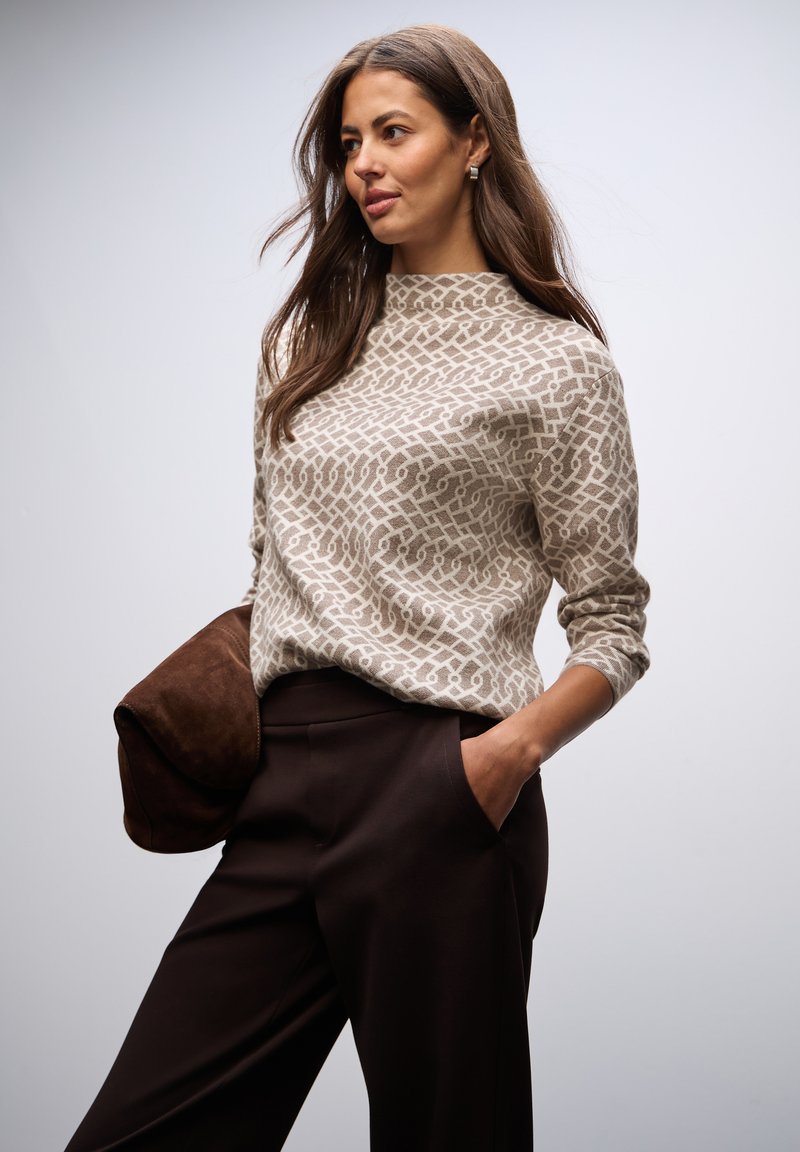 Beżowy sweter w geometryczny wzór, z wysokim kołnierzem i długimi rękawami, zestawiony z ciemno-brązowymi spodniami i brązową torebką z zamszu.