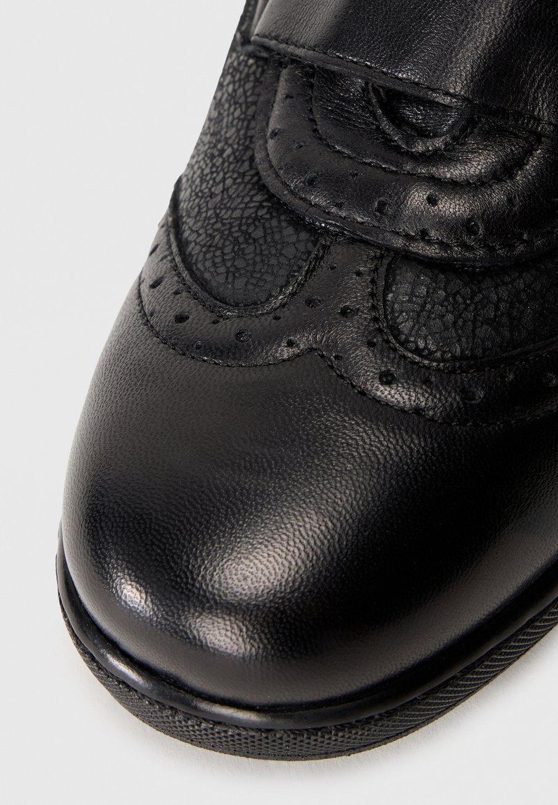 Scarpa di pelle nera con finitura liscia e accento testurizzato. Presenta una punta arrotondata, perforazioni decorative e una fascetta in velcro.