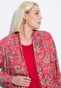 Punainen kuvioitu kimono, jossa on paisley- ja kukkakuosiset yksityiskohdat, kevyt kangas, pitkät hihat ja avonainen etuosa, joka on kerroksittain punaisen paidan päällä.