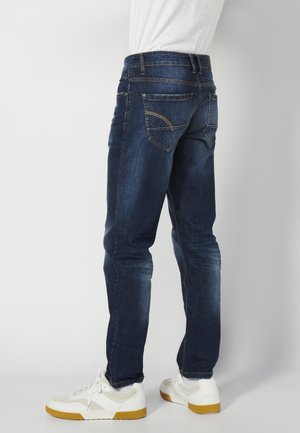 Férfi szabott sötétkék farmernadrág, vászon anyagból, öt zsebbel és varrási részletekkel, enyhe kopással a combok mentén, fehér sportcipővel párosítva.