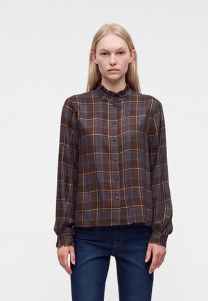 Femme portant un chemisier à carreaux marron et gris avec boutons, manches longues et col volanté, associé à un jean bleu foncé, debout sur fond blanc.