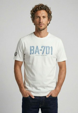 Homme aux cheveux bouclés portant un t-shirt blanc BA-701 et un jean foncé, debout les mains dans les poches devant un fond uni.