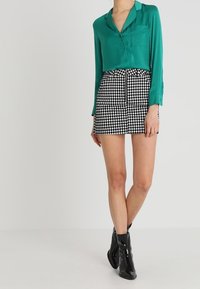Blusa in satin verde con tasca frontale, abbinata a una minigonna a scacchi bianchi e neri e stivaletti neri alla caviglia. Tessuto morbido, vestibilità su misura.