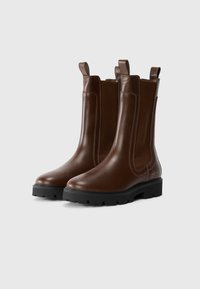 Une paire de bottes Chelsea en cuir marron foncé avec des semelles noires épaisses et des panneaux élastiques latéraux, présentée sur un fond clair uni.
