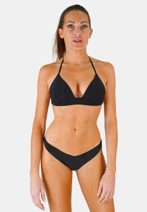 Femme aux cheveux bruns portant un haut de bikini noir à col halter et un bas de bikini taille haute assorti, debout devant un fond clair uni.