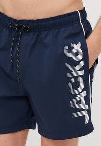 Marinefarbene Schwimmshorts mit elastischem Bund und schwarzem Kordelzug. Mit großem weißen Schriftzug auf dem linken Bein und seitlicher Tasche.