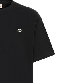 T-shirt noir à manches courtes avec un petit patch logo ovale blanc et noir sur la poitrine gauche avec les lettres S et D.