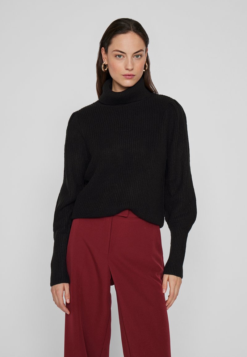 Pulover negru tricotat cu guler înalt și mâneci lungi, ușor bufante, asortat cu pantaloni largi roșii închis.