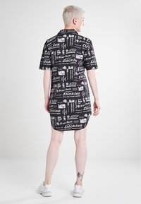 Abito a camicia nero con una stampa grafica bianca ripetitiva che include testo e immagini. Maniche corte, colletto e lunghezza fino a metà coscia.