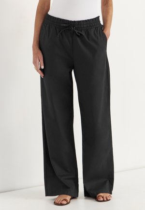 Persoon draagt losse zwarte broek met trekkoord, wit mouwloos topje en bruine sandalen, staand tegen een effen lichte achtergrond.