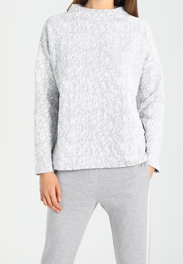 Femme portant un pull à manches longues gris clair texturé et un pantalon de survêtement gris clair avec une bande blanche sur le côté.