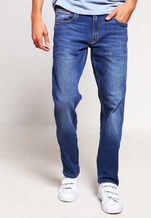 Man draagt blauwe denim jeans en witte klittenbandsneakers, staat met één hand in de zak tegen een effen achtergrond.