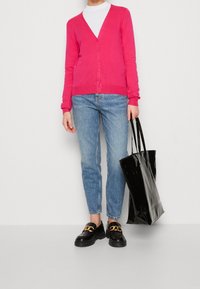 Różowy sweter na drutach z dekoltem w serek i guzikami, zestawiony z jasnoniebieskimi dżinsami, czarnymi loafersami z złotym łańcuszkiem oraz błyszczącą czarną torbą typu tote.