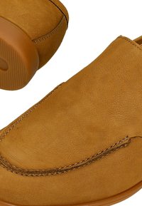 Scarpe slip-on in suede marrone con punta arrotondata e cuciture decorative. La suola è testurizzata, presenta un design piatto senza hardware visibile.